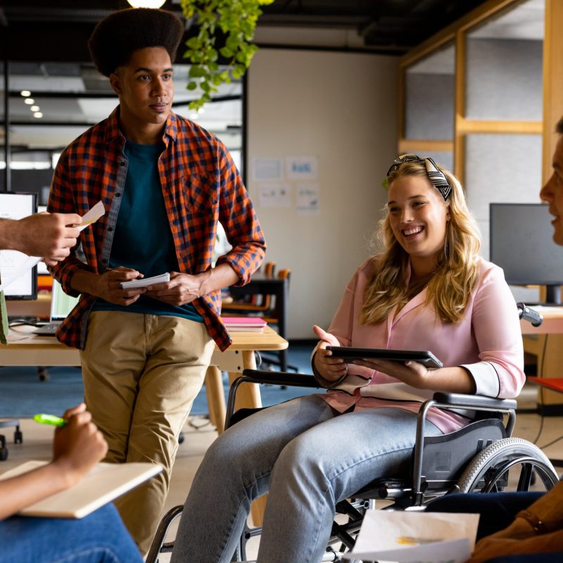 Happy diverse male and female colleagues in discussion using tablet in casual office meeting. Casual office, teamwork, disability, inclusivity, business and work, unaltered.